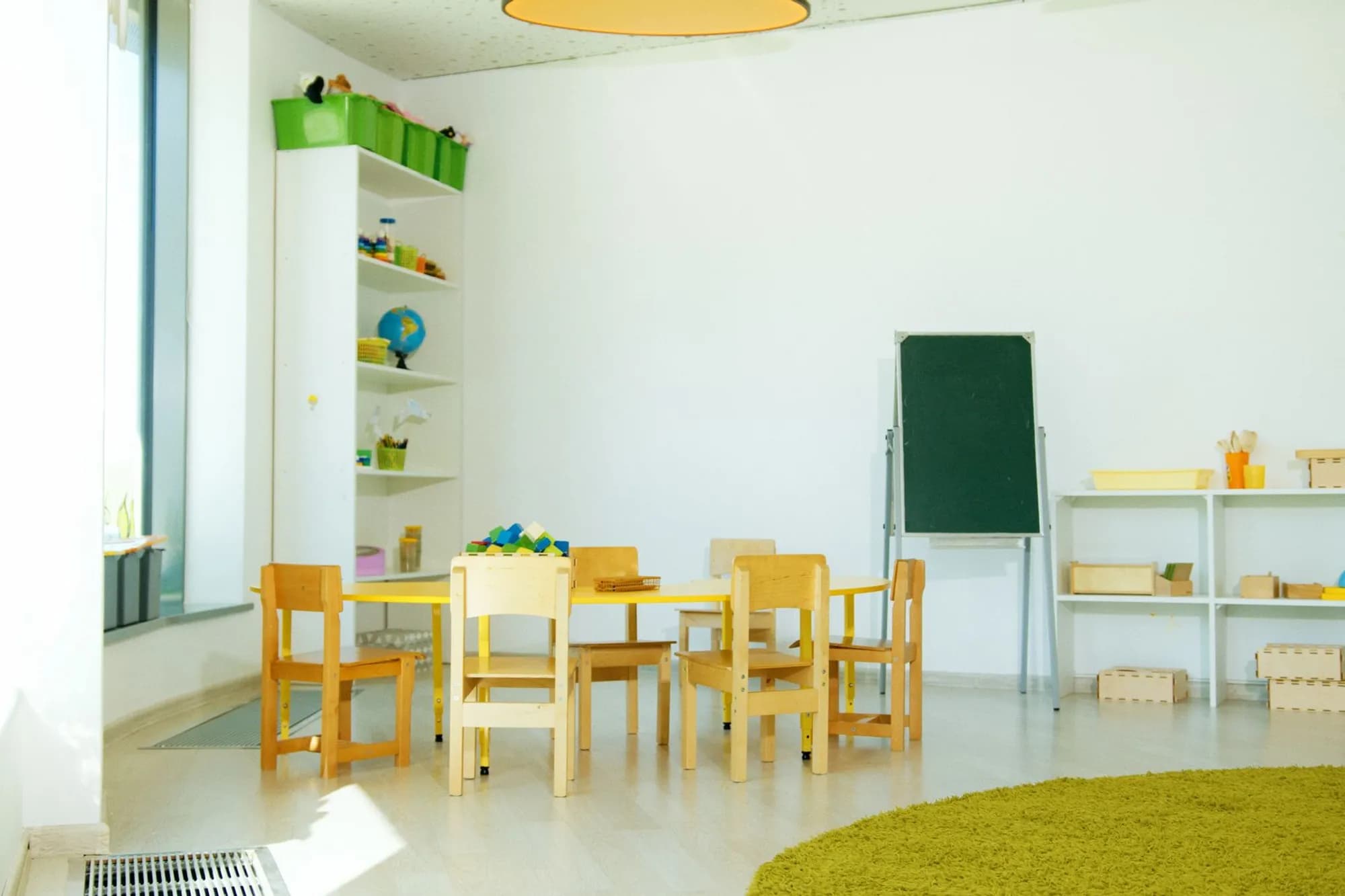 Leerer Gruppenraum mit Kindertisch, kleinen Stühlen, Tafel und Regalen